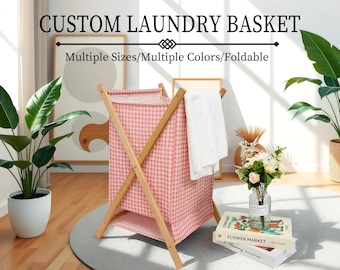 Puede incluir: Una cesta de ropa de color rosa y blanco a cuadros con un marco de madera. La cesta es plegable y tiene una toalla blanca colgada en el lateral. El texto "CUSTOM LAUNDRY BASKET" y "Multiple Sizes/Multiple Colors/Foldable" se muestra encima de la cesta.