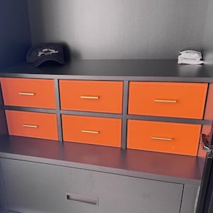 May include: A black cabinet with six orange drawers and a black drawer below. The cabinet has a black top with a black baseball cap with the text "NEW YORK" on it.