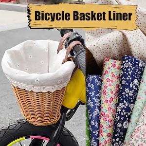 Op de afbeelding: Een rieten fietstas met een witte stoffen voering met een witte kanten rand. De tas is bevestigd aan een gele fiets. De afbeelding toont ook verschillende stoffen met bloemmotieven.