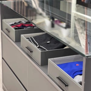 May include: A gray dresser with four drawers and a glass top. The top drawer is open and contains folded clothing. The second drawer is open and contains a sweater. The third drawer is open and contains a blue shirt. The drawers have black handles.