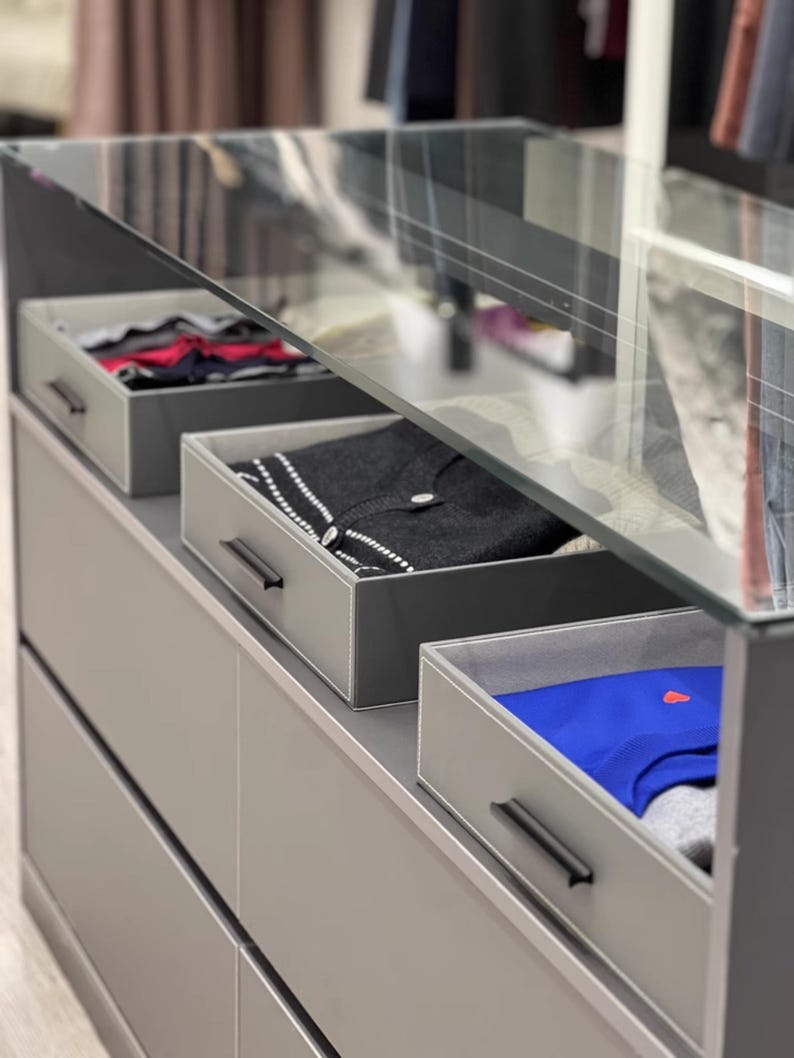 May include: A gray dresser with four drawers and a glass top. The top drawer is open and contains folded clothing. The second drawer is open and contains a sweater. The third drawer is open and contains a blue shirt. The drawers have black handles.