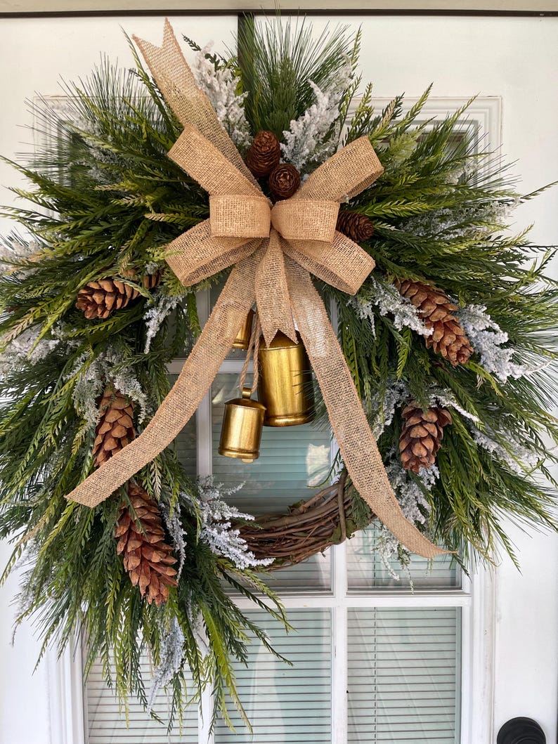 May include: A 24-inch artificial Christmas wreath features a rustic design.  The wreath is predominantly green with frosted pine, cedar, and juniper accents.  Brown pinecones and gold bells are interspersed throughout the greenery. A burlap bow adds a natural touch. This festive wreath is perfect for a front door or wall decor.
