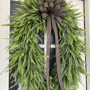 May include: A full, green fern wreath adorned with a brown and white striped bow and streamers, hanging on a white door. The wreath's dense fern leaves create a vibrant, textured appearance. The bow adds a touch of elegance to the natural, welcoming design.