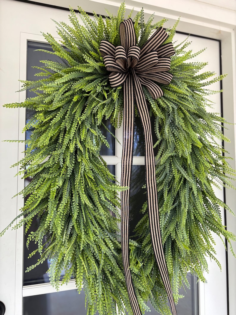 May include: A full, green fern wreath adorned with a brown and white striped bow and streamers, hanging on a white door. The wreath's dense fern leaves create a vibrant, textured appearance. The bow adds a touch of elegance to the natural, welcoming design.