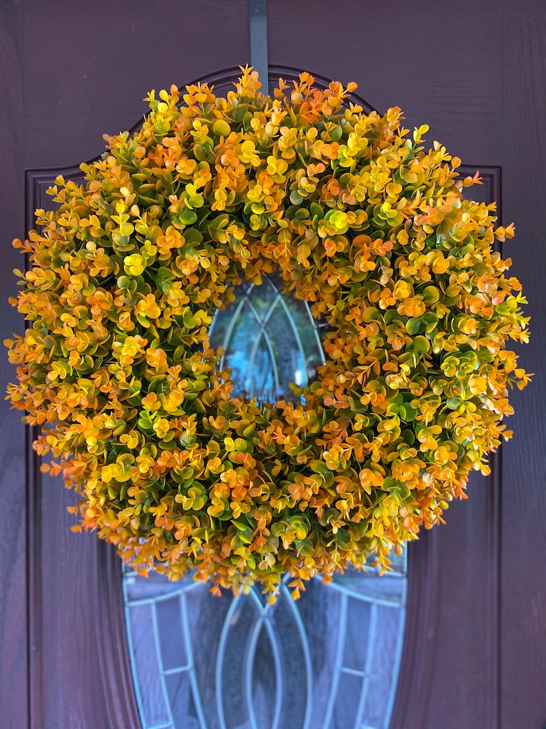 May include: A yellow and green artificial wreath made of small leaves. The wreath is hanging on a brown door with a glass window pane.