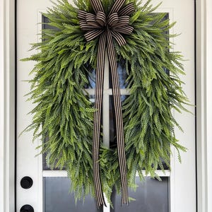 May include: A green fern wreath with a brown and cream striped bow and streamers hangs on a white door. The wreath is full and lush, with a natural, textured appearance. The door has black hardware.