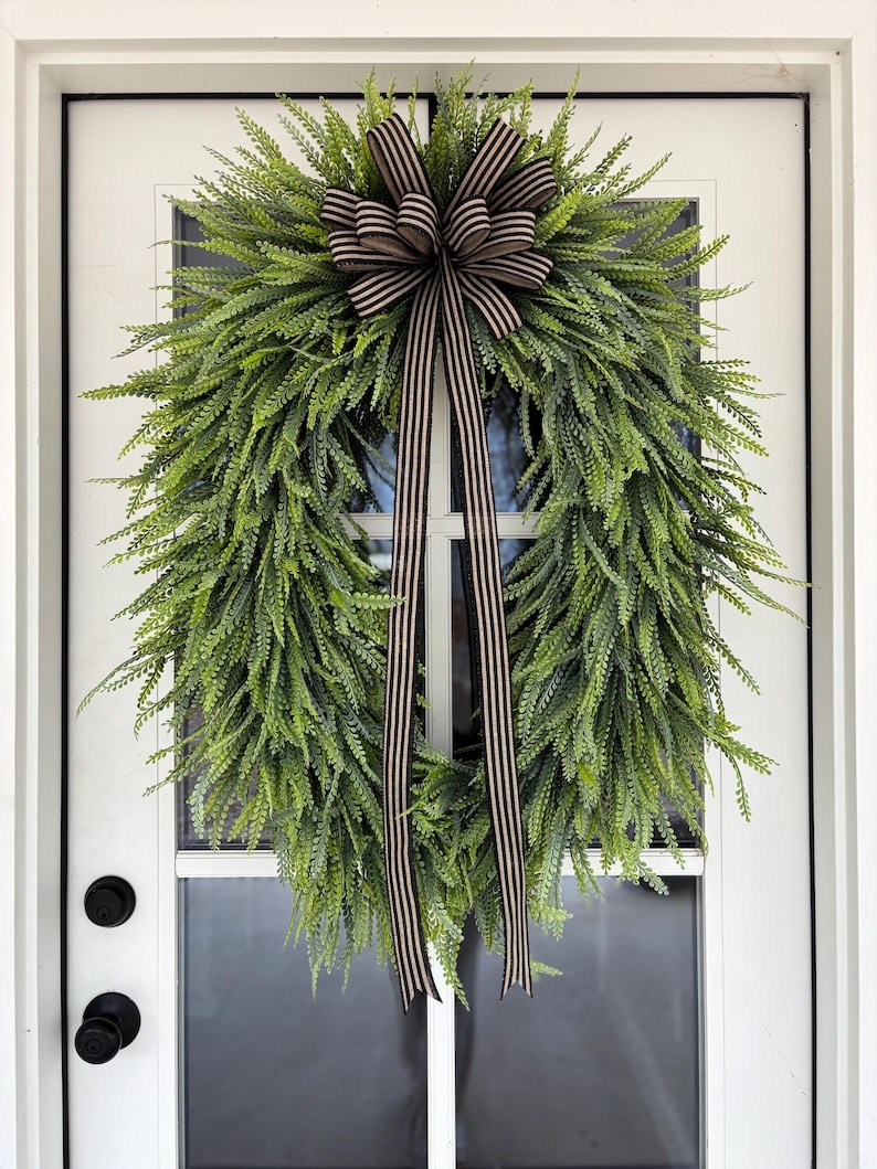 May include: A green fern wreath with a brown and cream striped bow and streamers hangs on a white door. The wreath is full and lush, with a natural, textured appearance. The door has black hardware.
