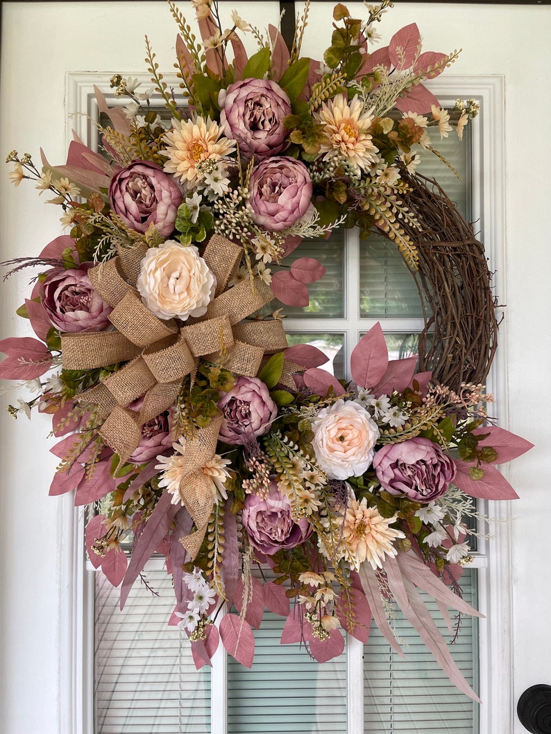 May include: A pink and cream floral wreath with a burlap bow. The wreath is made of artificial flowers and greenery and is perfect for a spring or summer door decoration.