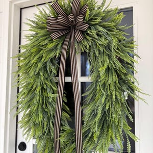 May include: A green fern wreath with a brown and cream striped bow and streamers hangs on a white door. The wreath is full and lush, with long, delicate leaves. The bow is centered at the top, adding a touch of elegance.