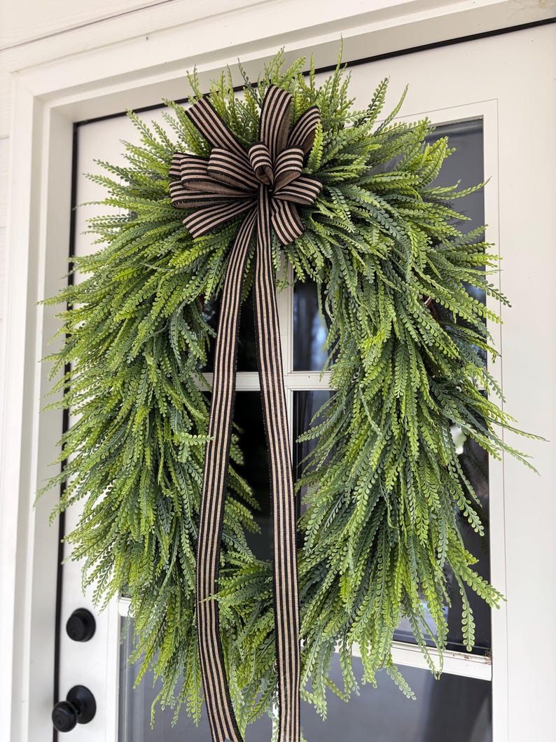 May include: A green fern wreath with a brown and cream striped bow and streamers hangs on a white door. The wreath is full and lush, with long, delicate leaves. The bow is centered at the top, adding a touch of elegance.