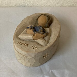 Puede incluir: Una pequeña caja de cerámica de forma ovalada con un diseño esculpido de una mujer sosteniendo un ramo de flores. La caja está pintada en tonos beige y marrón y tiene las palabras "Thank You" inscritas en la parte delantera.