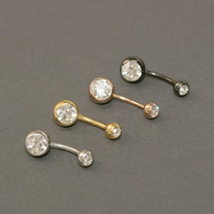 May include: Four curved belly button rings with clear, round gemstones. The rings are in silver, gold, rose gold, and black metal finishes. The gemstones are set in a bezel setting, and the smaller gem is at the bottom of the curve.
