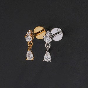Peut inclure: Deux boucles d'oreilles à clous, une dorée et une argentée, chacune ornée d'une petite gemme ronde et d'une gemme en forme de poire pendante. Les boucles d'oreilles sont présentées sur un fond noir, mettant en valeur leur éclat. La boucle d'oreille dorée a une tige dorée et la boucle d'oreille argentée a une tige argentée.