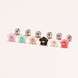 May include: Six flower-shaped stud earrings in various colors: lavender, mint green, pink, black, white, and red. Each earring has a silver post and a small gold-colored center. The earrings are displayed on a light pink surface.