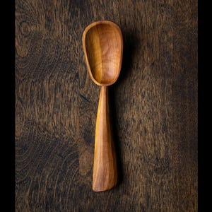 May include: A handcrafted wooden spoon with a rounded bowl and a long, tapered handle. The spoon is made of light brown wood and sits on a dark brown wooden surface. The spoon's design suggests it is for cooking or serving.