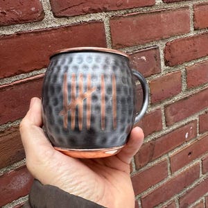 May include: A copper-rimmed Moscow Mule mug with a textured, dark gray finish. The mug features a copper design on the front. The handle is silver. The mug is held in front of a red brick wall.
