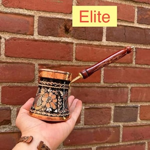 May include: A copper coffee pot with a black exterior featuring an intricate orange and white floral pattern. The pot has a metallic rim and a long, dark brown wooden handle with decorative carvings. The word Elite is printed in red on a yellow box.