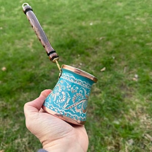 May include: A turquoise and copper coffee pot with an ornate floral design. The pot has a wooden handle and is held in a hand against a green grass background. The pot is likely used for making Turkish or Greek coffee.