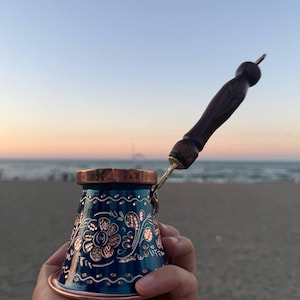 May include: A metallic blue coffee pot with a copper rim and base, featuring intricate hand-engraved floral patterns that reveal the copper metal underneath. The pot includes a long, dark brown wooden handle with a carved, textured grip.