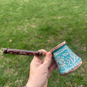 May include: A turquoise and copper coffee pot with a long, ornate wooden handle. The pot features a floral design and a copper rim. The handle is dark brown with a decorative pattern. The pot is held against a background of green grass.