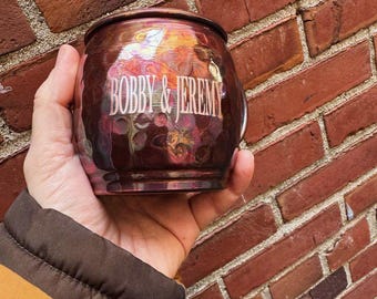 May include: A round, metallic mug with a dark, iridescent finish featuring swirling hues of purple, gold, and bronze. The text BOBBY & JEREMY is printed in a light, serif font across the center of the mug, which has a smooth, reflective surface.