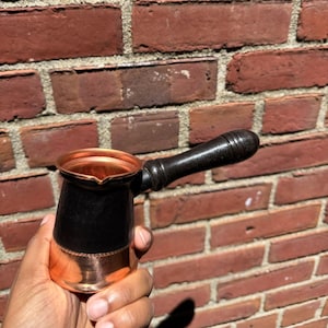 以下が含まれることがあります： 木製のハンドルが付いた銅と黒のコーヒーポット。ポットは、光沢のある銅の内側とベース、黒の外側を備えています。ハンドルは濃い茶色で湾曲しています。ポットは赤いレンガの壁の前に保持されています。