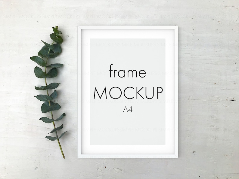 May include: White frame mockup with the text "frame MOCKUP A4" on a white wood background with a sprig of eucalyptus leaves to the left of the frame.