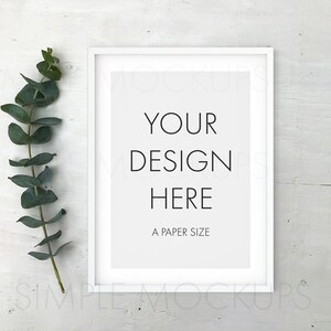 May include: White frame mockup with a white mat and the text "YOUR DESIGN HERE A PAPER SIZE" on a white wood background with a sprig of eucalyptus leaves to the left of the frame.