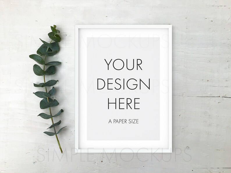 May include: White frame mockup with a white mat and the text "YOUR DESIGN HERE A PAPER SIZE" on a white wood background with a sprig of eucalyptus leaves to the left of the frame.