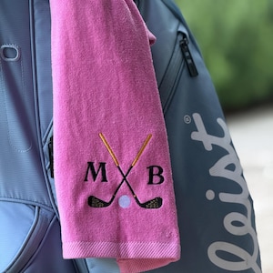 May include: A pink golf towel with embroidered golf clubs, a golf ball, and the letters M, X, and B. The towel is hanging on a blue golf bag.