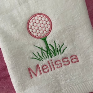 May include: White golf towel embroidered with a pink golf ball on a tee and green grass. The name "Melissa" is embroidered in pink below the golf ball design. The towel is folded and resting on a pink towel.