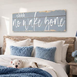 May include: A rectangular wooden wall sign with a distressed blue finish and white script text that reads shhh... no wake zone. The sign is mounted horizontally above a bed featuring a chunky blue knit throw blanket and two blue pillows.