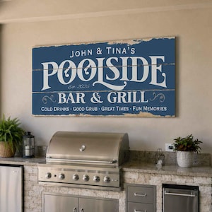 May include: A rectangular blue wooden sign with white text reading John & Tina's POOLSIDE BAR & GRILL Est. 2026, featuring the additional text Cold Drinks, Good Grub, Great Times, Fun Memories, displayed on a wall above an outdoor kitchen area.