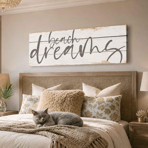 May include: A rectangular wall sign with a distressed white finish and horizontal wood grain texture. The text "beach dreams" is printed in a dark gray, cursive script across the center of the sign, spanning the width of the board.