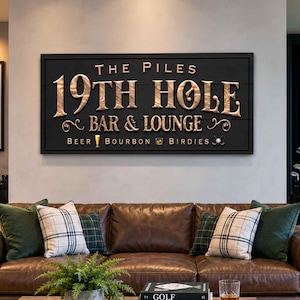 May include: A rectangular black wooden sign with raised gold lettering that reads THE PILES 19TH HOLE BAR & LOUNGE, with icons of a beer glass, a glass of bourbon, and a golf ball. The sign is mounted on a wall above a brown leather sofa.