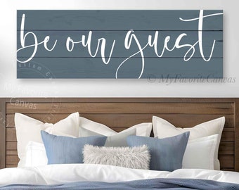 May include: A blue wooden sign with the words "be our guest" in white script is displayed above a bed. The bed is made with white linens, blue pillows, and a blue blanket. A wooden headboard and a bench complete the bedroom decor.