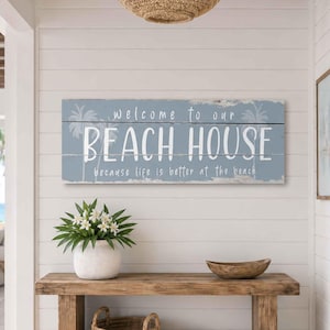 May include: A rectangular wooden wall sign with a distressed blue finish and white text that reads "welcome to our BEACH HOUSE because life is better at the beach" with faint white palm tree silhouettes in the background.