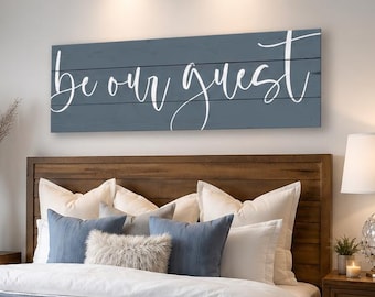 Puede incluir: Un letrero de pared rectangular de madera azul con el texto cursivo blanco "be our guest" montado sobre una estructura de cama de madera con ropa de cama blanca, una manta azul, un banco beige capitoné y dos mesitas de noche de madera con lámparas.