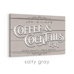 May include: A rectangular sign with the words "Coffee & Cocktails Bar" in white, raised lettering. The sign is light gray with horizontal wood grain lines. The text "Lori & Nick's" and "Open Daily - Self Serve" are also visible.