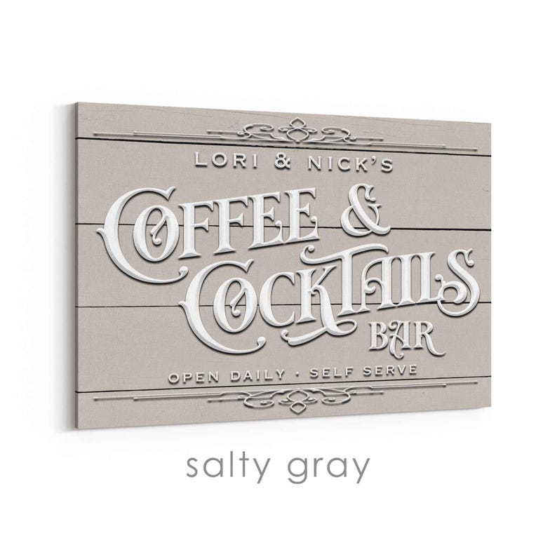 May include: A rectangular sign with the words "Coffee & Cocktails Bar" in white, raised lettering. The sign is light gray with horizontal wood grain lines. The text "Lori & Nick's" and "Open Daily - Self Serve" are also visible.
