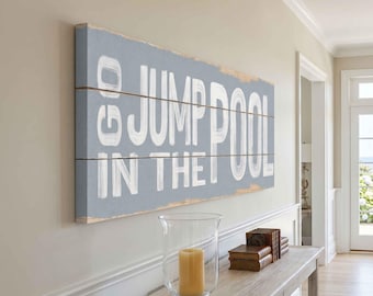 May include: A rectangular wooden sign with the words "GO JUMP IN THE POOL" in white lettering. The sign is painted light blue with a distressed look and is mounted on a wall. A candle in a glass holder sits on a wooden table below.