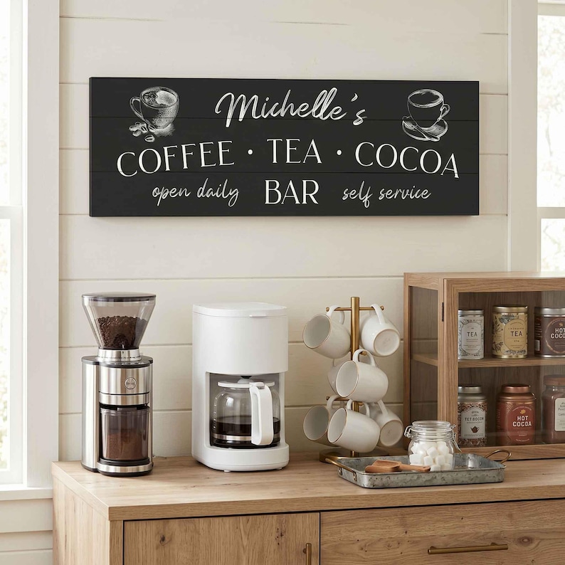 May include: A black wooden sign with white text reading "Michelle's COFFEE &bull; TEA &bull; COCOA open daily BAR self service" hangs above a coffee station. A coffee grinder, coffee maker, and mug rack are on a wooden counter.