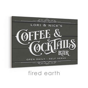 May include: A dark gray wooden sign with white lettering that reads "LORI & NICK'S COFFEE & COCKTAILS BAR." The sign also includes the text "OPEN DAILY - SELF SERVE" and decorative flourishes.