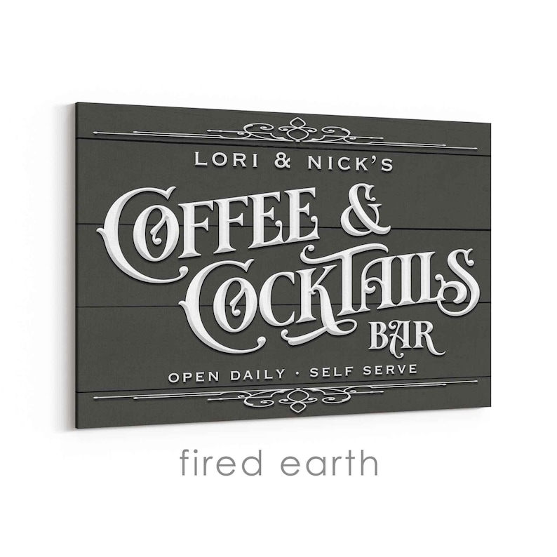 May include: A dark gray wooden sign with white lettering that reads "LORI & NICK'S COFFEE & COCKTAILS BAR." The sign also includes the text "OPEN DAILY - SELF SERVE" and decorative flourishes.