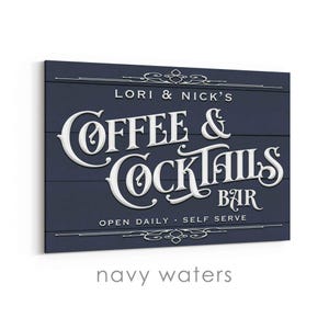 May include: Navy blue wooden sign with white lettering that reads "Lori & Nick's Coffee & Cocktails Bar." The sign also includes the text "Open Daily - Self Serve" and decorative flourishes.