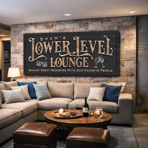 May include: A black wooden sign with gold lettering that reads "Shaw's Lower Level Lounge." The sign includes illustrations of glasses and text that says "Making Great Memories With Our Favorite People." The sign hangs above a beige sectional sofa.