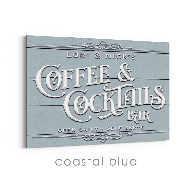 May include: A coastal blue wooden sign with white raised lettering that reads "Lori & Nick's Coffee & Cocktails Bar". The sign also includes the text "Open Daily - Self Serve" and decorative flourishes.