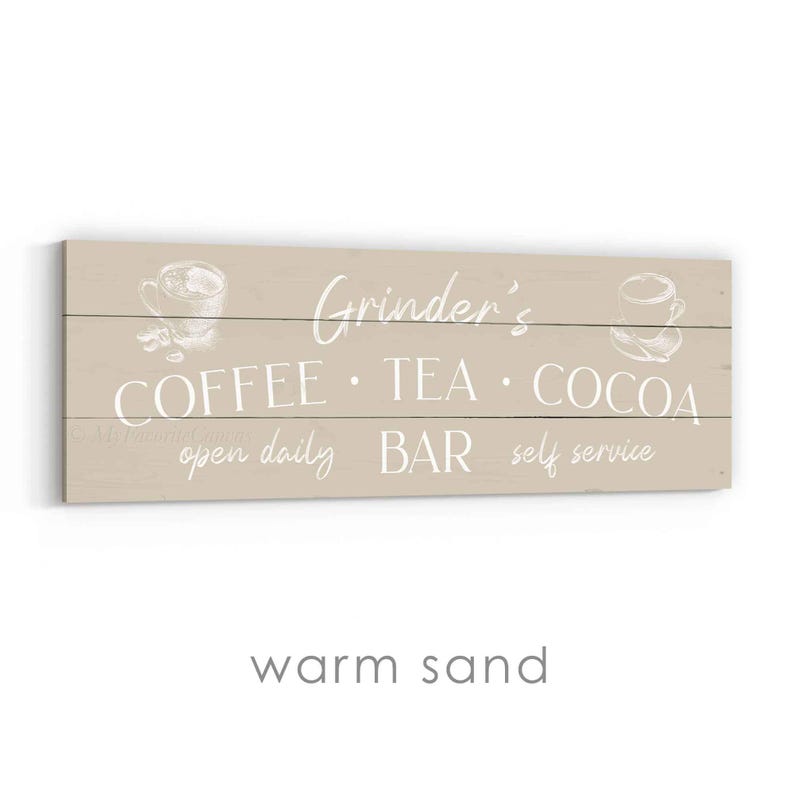 May include: A warm sand-coloured wooden sign with white text that reads "Grinder's COFFEE &bull; TEA &bull; COCOA BAR open daily self service." The sign has sketched images of coffee cups on each side.