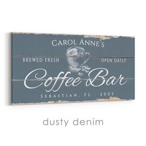 Pode incluir: Uma placa retangular com aspecto envelhecido, com fundo azul denim empoeirado. A placa diz "Carol Anne's Coffee Bar" em escrita branca, com uma ilustra&ccedil;&atilde;o de x&iacute;cara de caf&eacute;. Texto adicional: "Brewed Fresh", "Open Daily" e "Sebastian, FL 2003".
