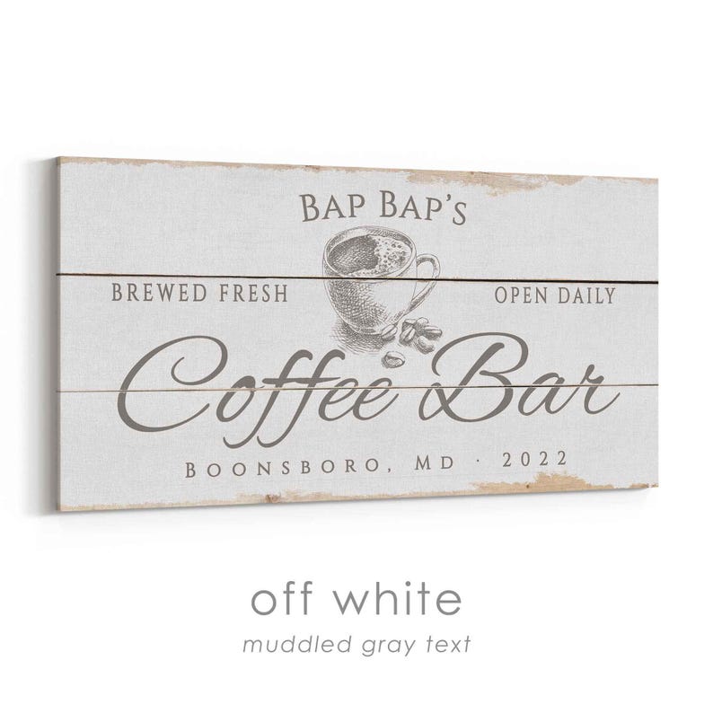 Pode incluir: Placa de madeira branca suja com texto cinza turvo. A placa diz "Bap Bap's Coffee Bar" com uma ilustra&ccedil;&atilde;o de x&iacute;cara de caf&eacute;. O texto adicional inclui "Brewed Fresh", "Open Daily", "Boonsboro, MD 2022".